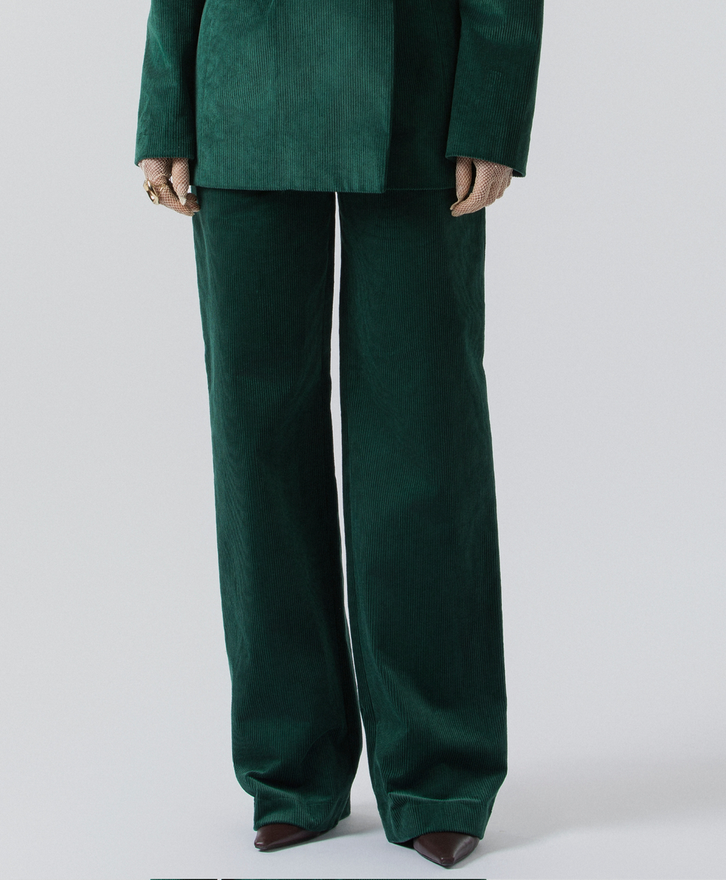 A person wearing a dark green velvet-like blazer and matching wide-leg trousers, shown from the shoulders down against a light gray studio background.