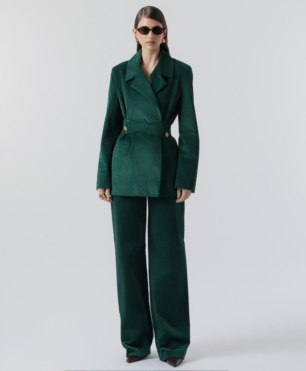 A model wearing a dark green velvet two-piece suit consisting of a belted blazer and matching wide-leg trousers, styled with dark sunglasses, against a plain light gray studio background.