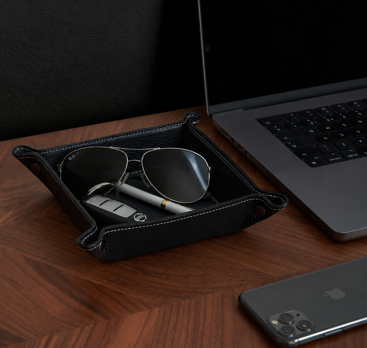 Black leather valet tray with sunglasses and car keys on a wooden desk next to a laptop and smartphone.
