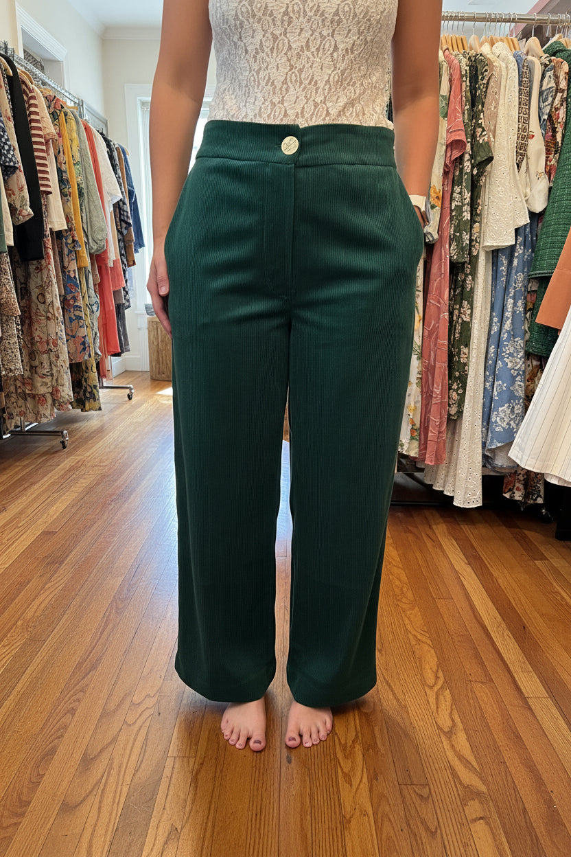 A person wearing dark green corduroy wide-leg cropped pants with a single gold button closure at the waist, standing in a clothing store.