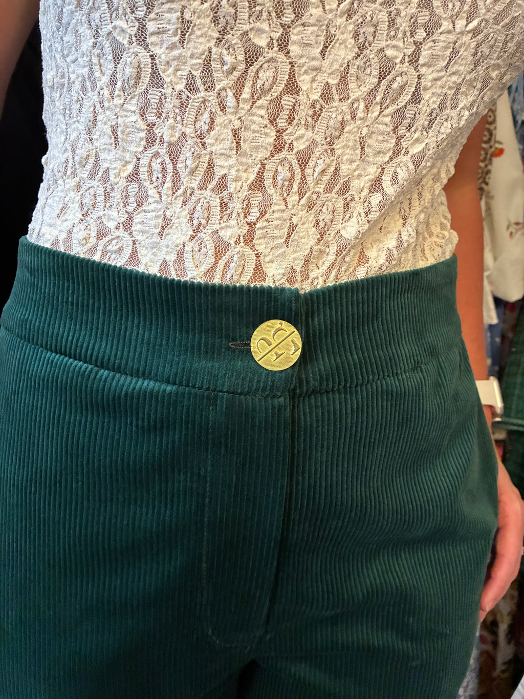 Close-up of a person wearing dark green corduroy pants with a gold button featuring a stylized 'T' logo, paired with a white lace top.