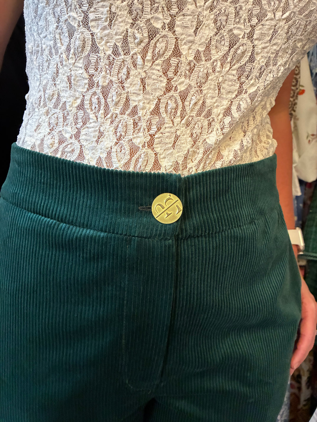 Close-up of a person wearing dark green corduroy pants with a gold button featuring a stylized 'T' logo, paired with a white lace top.